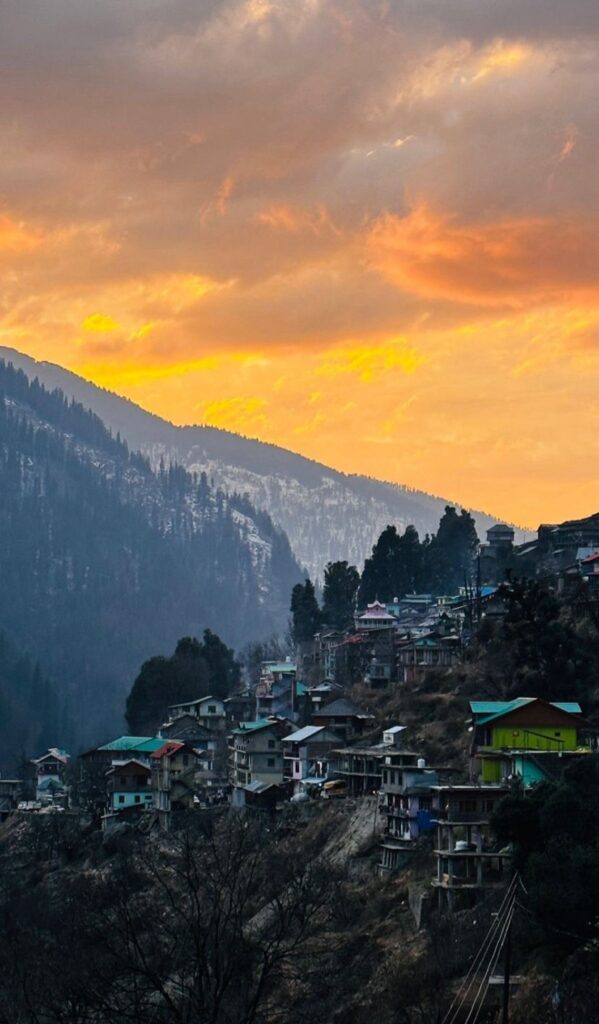 Colorful mountain village on hillside with snow-capped peaks and dramatic sunset sky